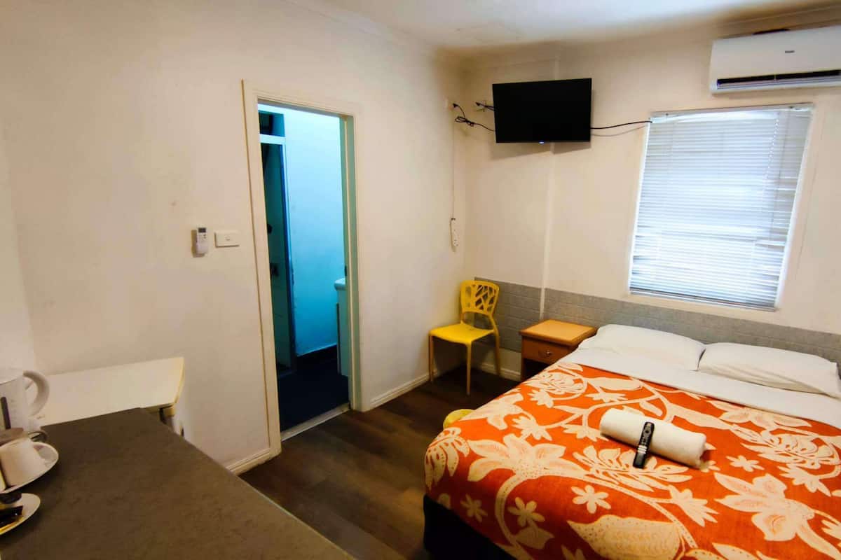 A small, well-lit room with a bed, a chair, a small table, a TV mounted on the wall, and an air conditioning unit.