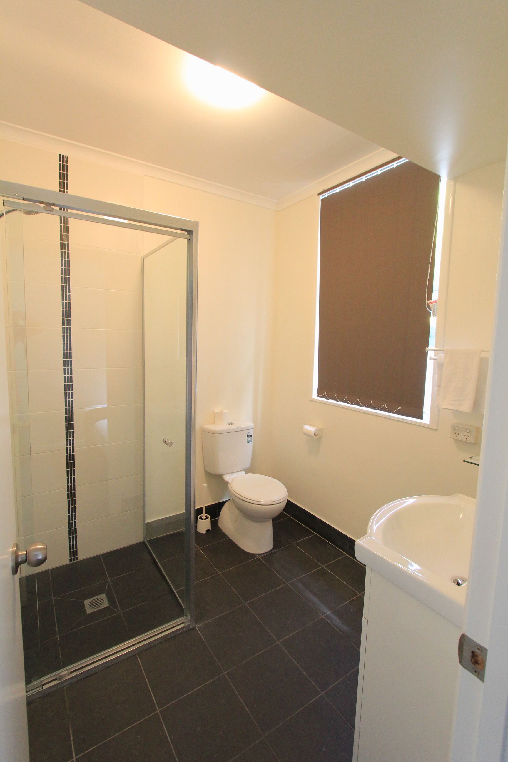 A bathroom with a glass shower enclosure, a toilet, a sink, and a window with blinds.