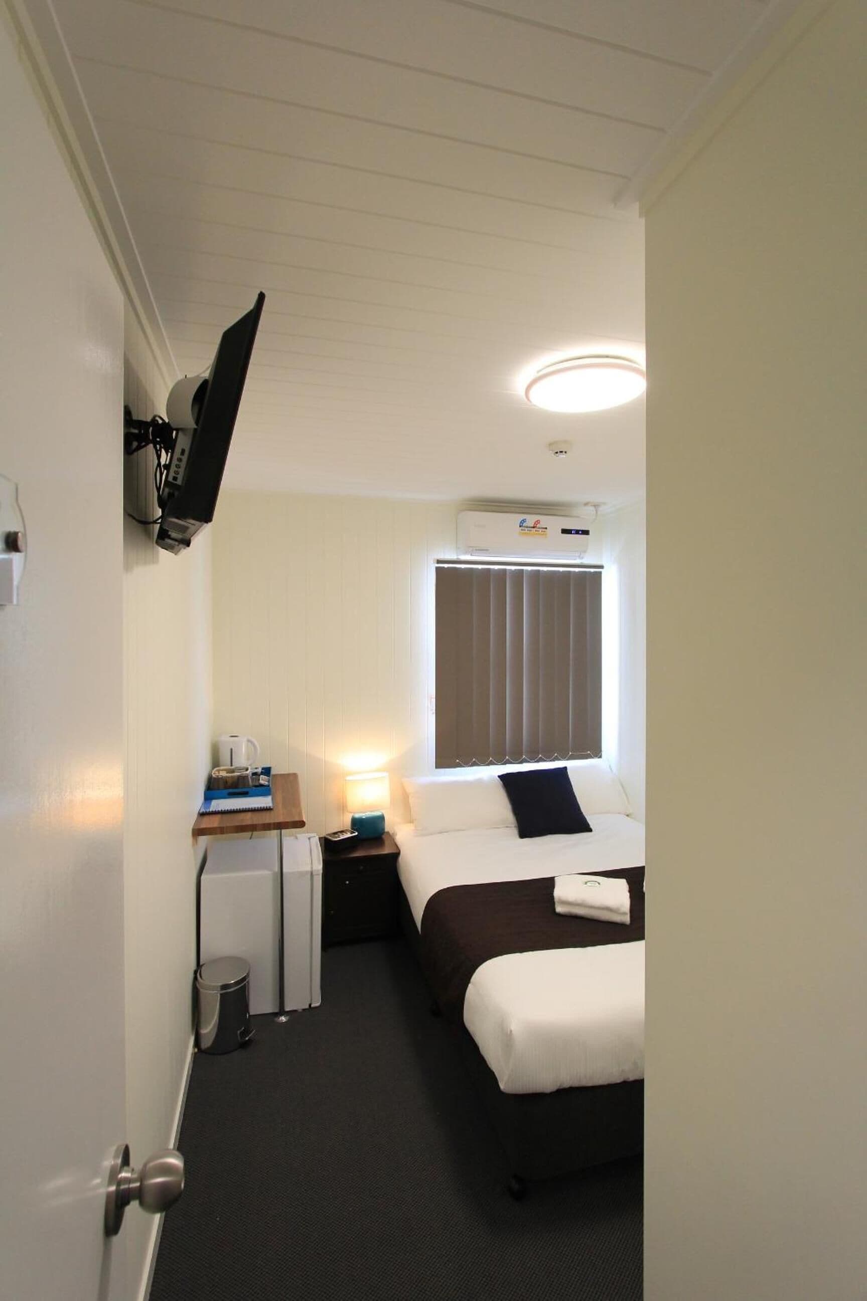 Habitación de hotel con cama, escritorio con lámpara, ventana con persianas, unidad de aire acondicionado y televisor montado en el techo.