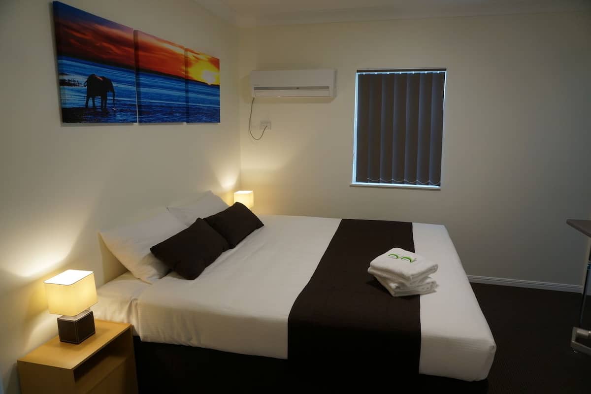 A hotel room with a bed, a nightstand with a lamp, an air conditioner, a window with blinds, and a painting of an elephant on the wall.