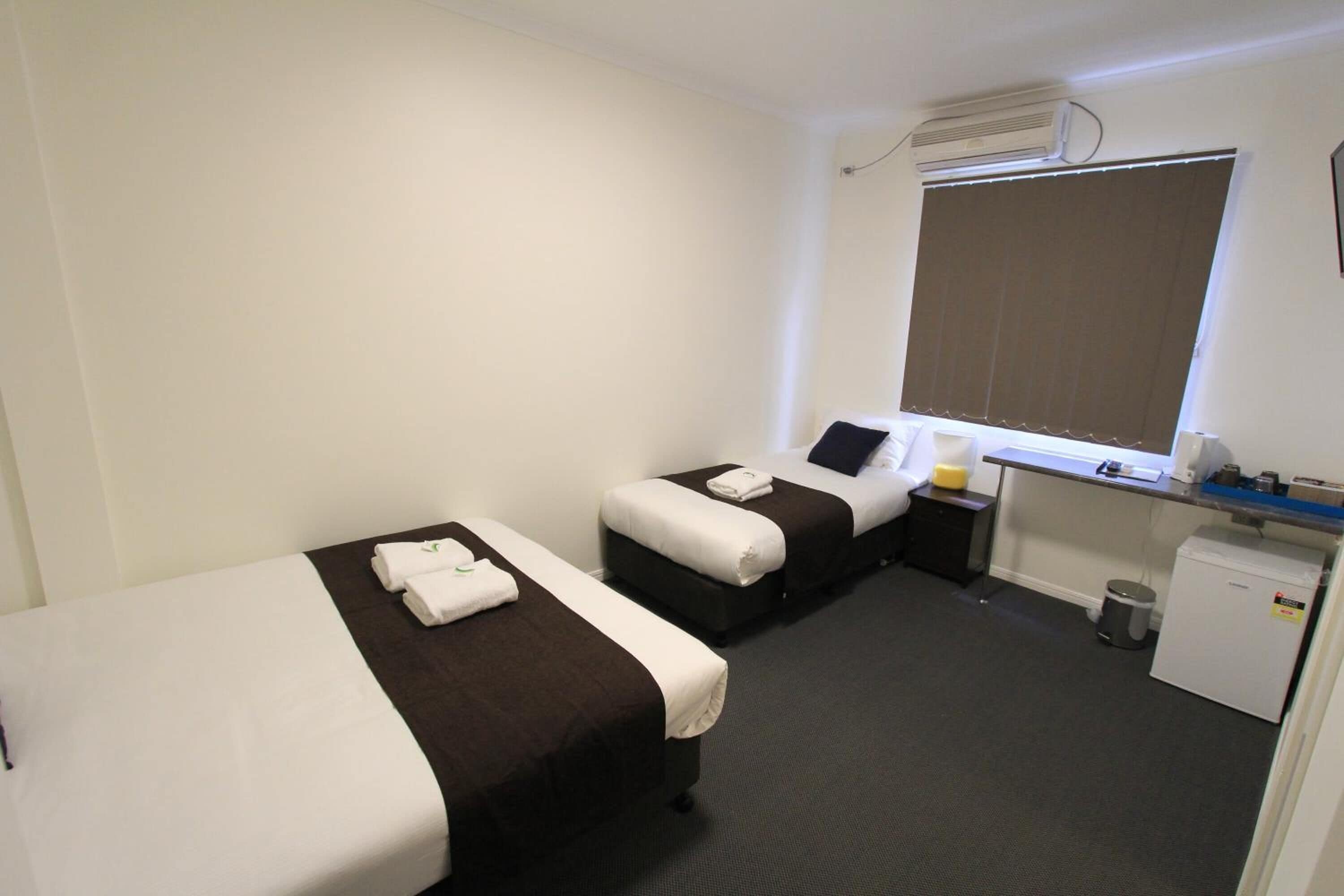 Ein Hotelzimmer mit zwei Einzelbetten, einem Schreibtisch mit Computer, einer kleinen Kühlbox und einem Fenster mit Jalousien.