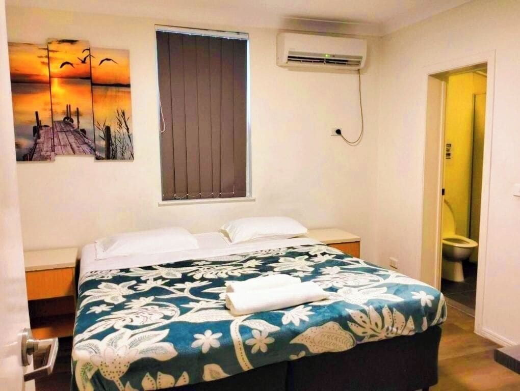 A bedroom with a bed, a window with blinds, an air conditioning unit, and a painting on the wall.