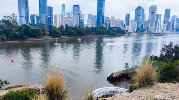A river with boats, a city skyline in the background, and a park with trees and grass in the foreground.