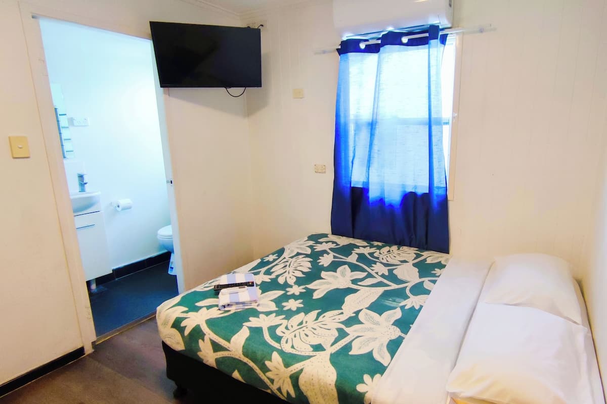 A hotel room with a bed, a TV mounted on the wall, a bathroom with a toilet and sink, and a window with blue curtains.