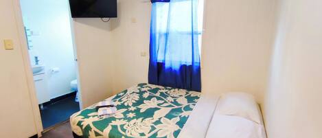 Habitación de hotel con cama, televisor montado en la pared, baño con inodoro y lavabo, y ventana con cortinas azules.