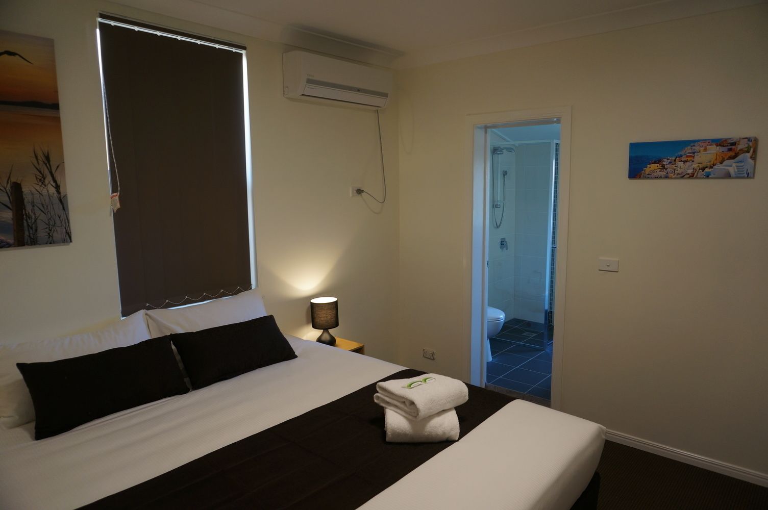 A bedroom with a bed, a nightstand, a lamp, a window with blinds, an air conditioning unit, a painting, and a bathroom door.