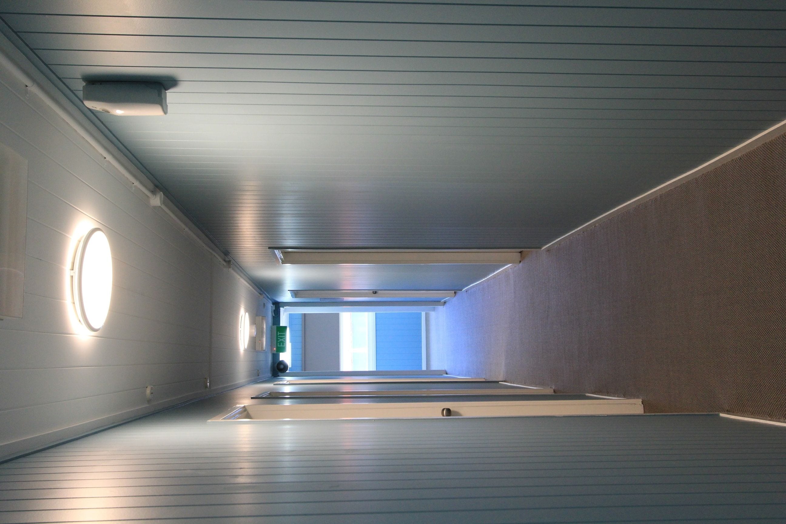 Un couloir avec un mur recouvert de miroirs, un luminaire circulaire et une porte au fond.