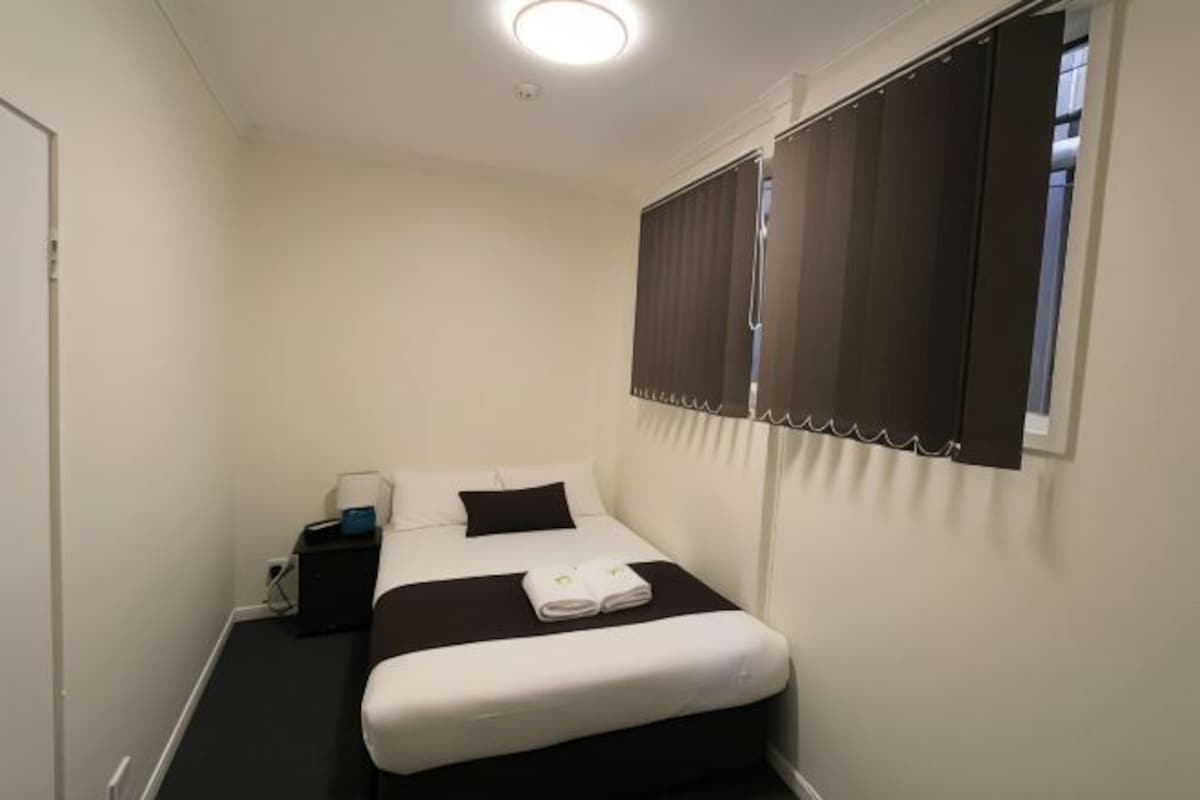 Une petite chambre à coucher, bien rangée, avec un lit simple, une table de chevet et une fenêtre avec des stores.