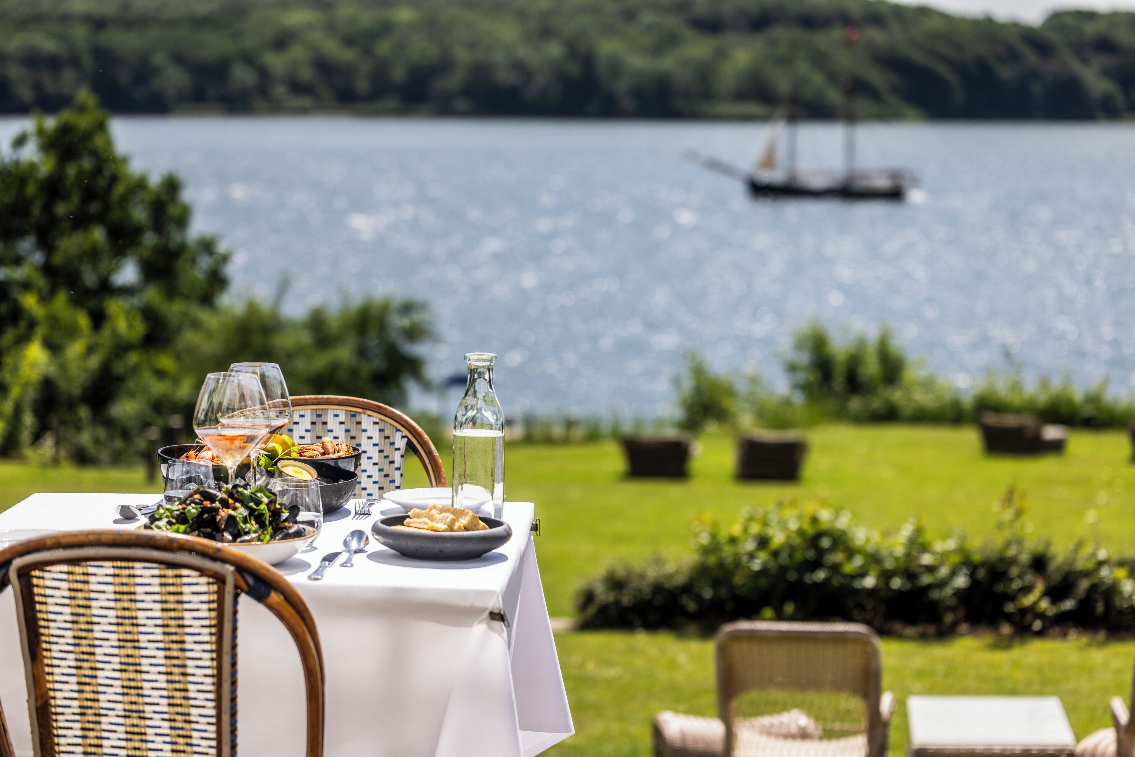 Mesa al aire libre con mariscos, copas de vino y una botella, con vista a un lago y follaje.