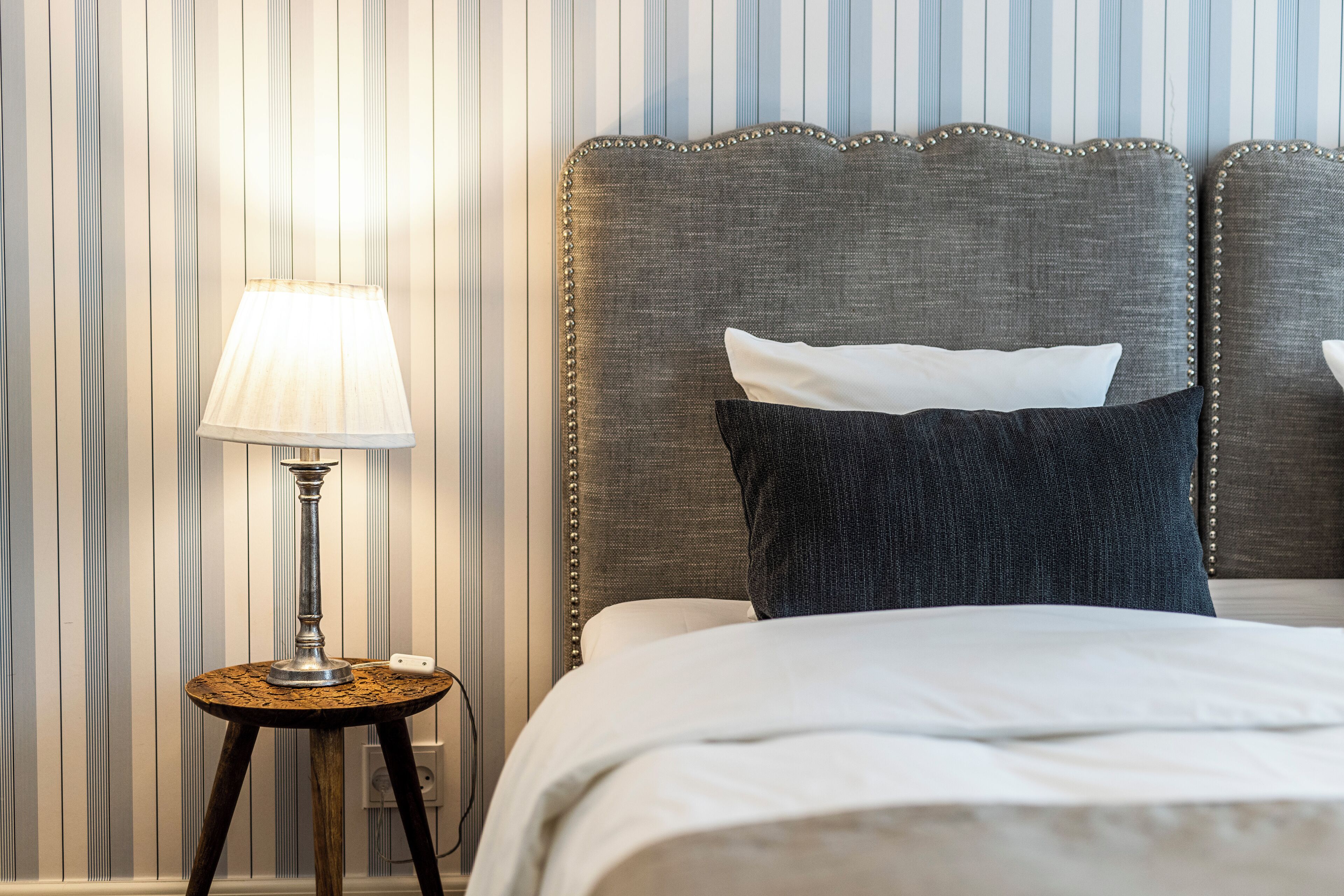 A neatly made bed with a grey upholstered headboard, a white pillow, and a dark pillow. A small wooden side table with a lamp is beside the bed.
