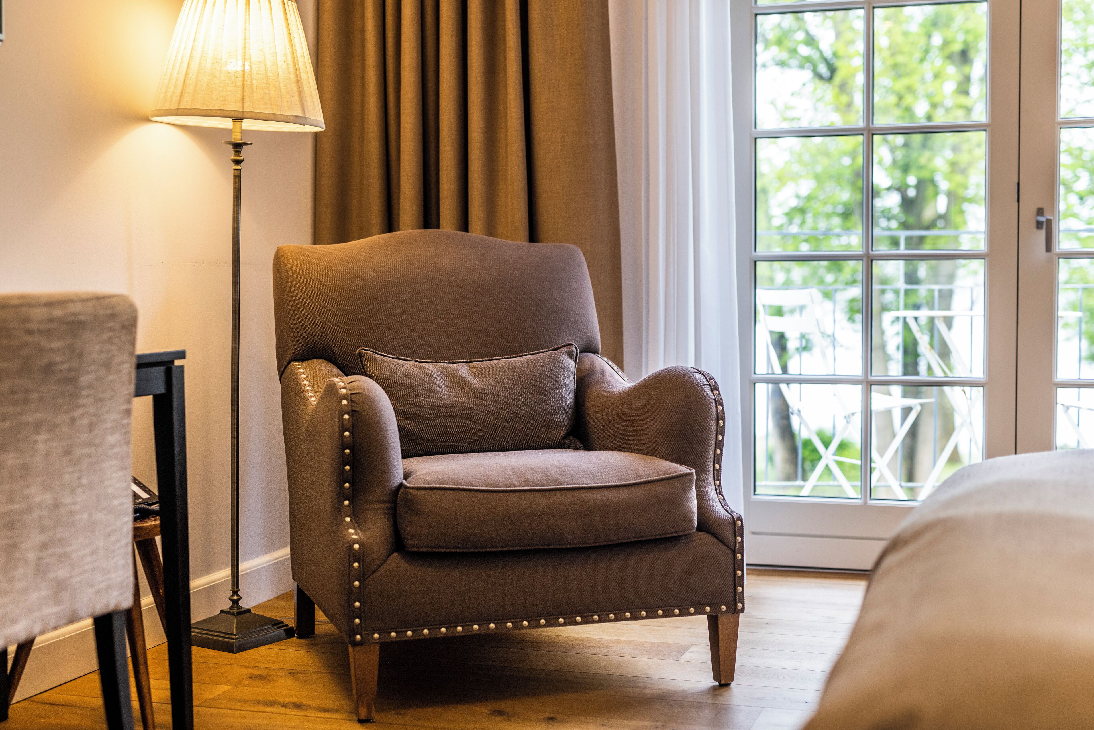Un fauteuil marron avec un coussin, un lampadaire et une vue sur la verdure par une fenêtre.