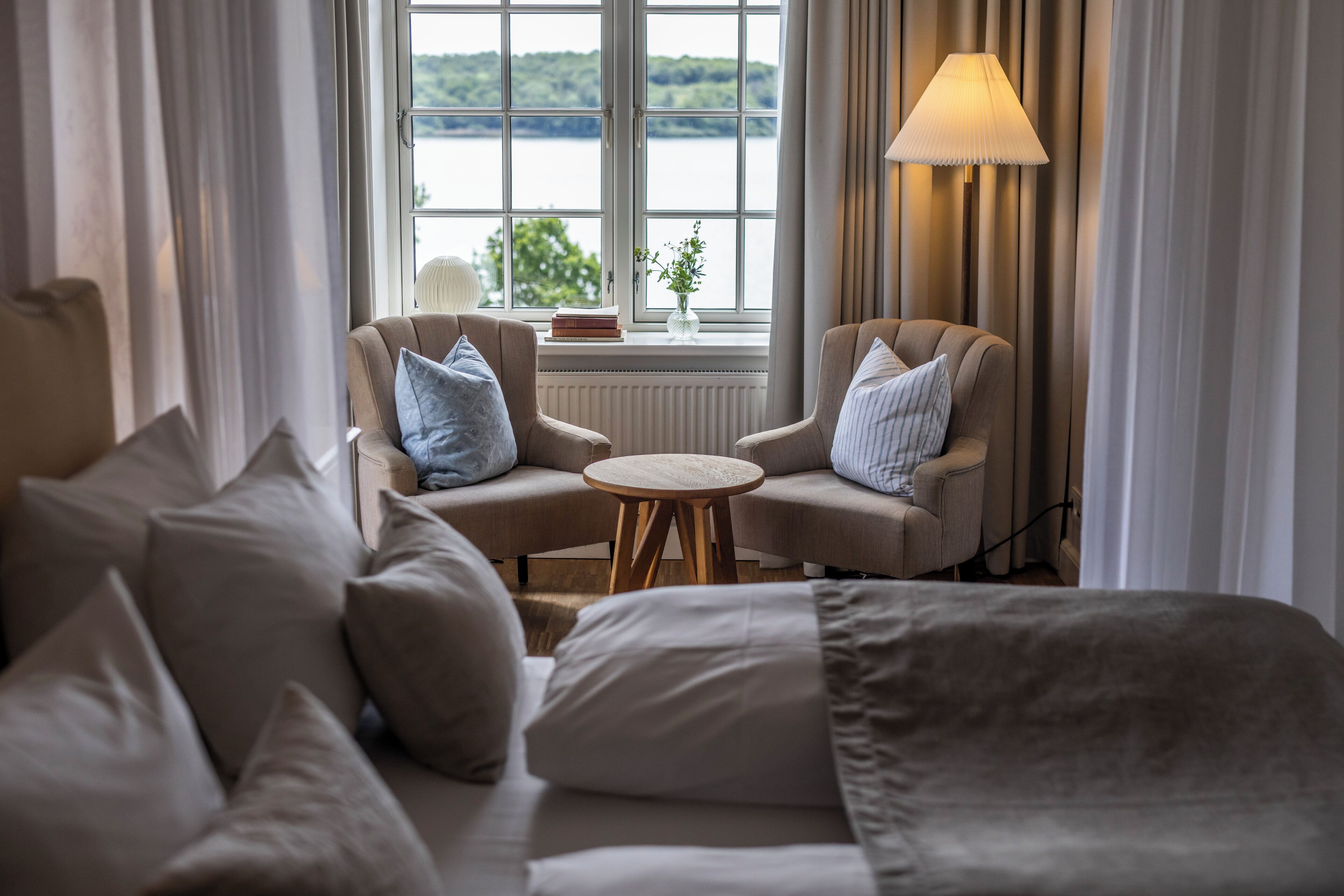 Habitación de hotel con sofá, dos sillones, una mesita redonda y vista a un lago a través de un ventanal grande.