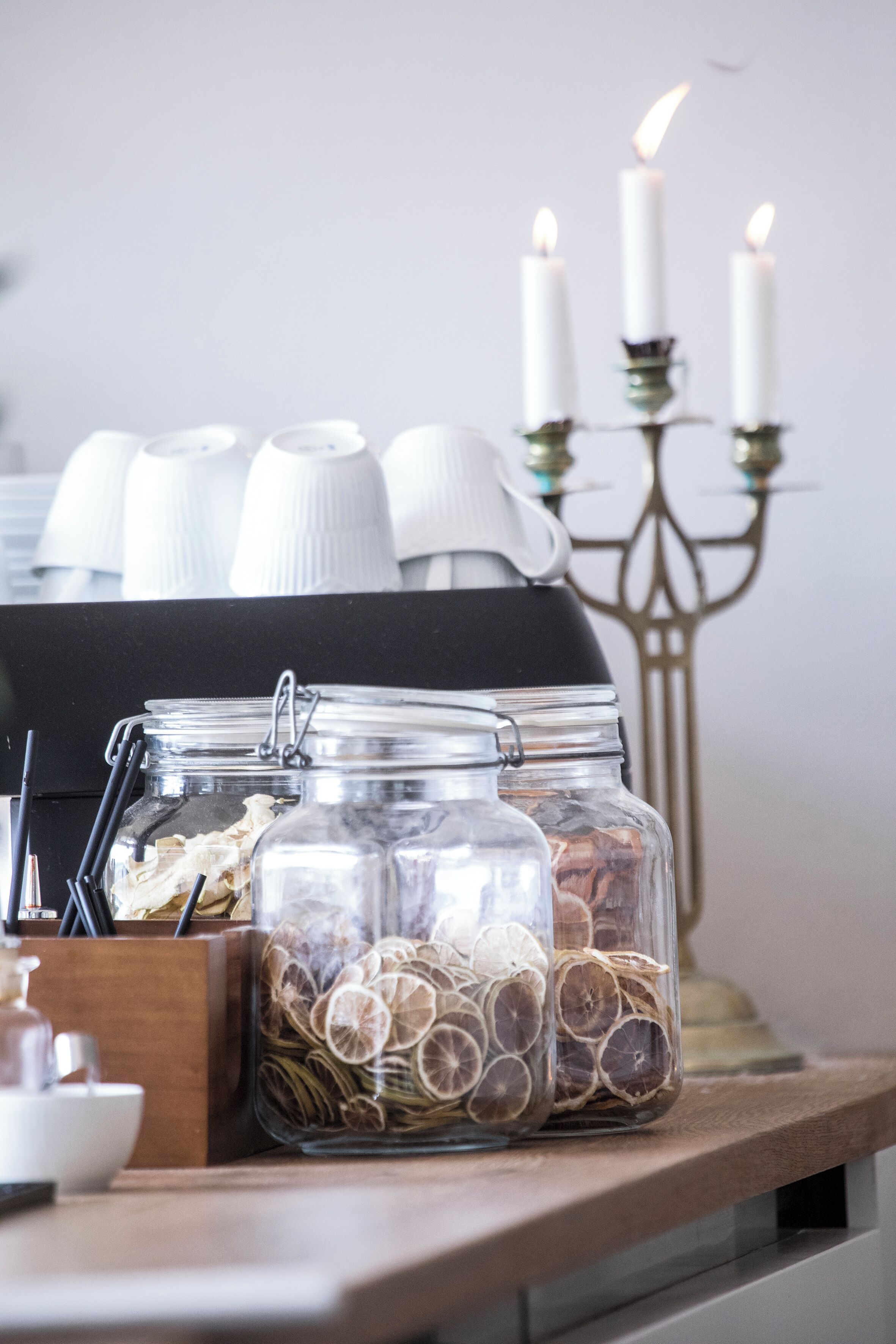Uma mesa com potes de vidro contendo fatias de cítricos secos, um castiçal com velas acesas e uma cafeteira.