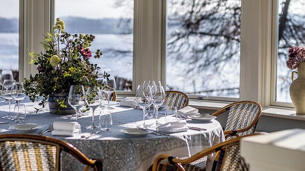 A dining room with a table set for six, surrounded by wicker chairs, a chandelier, and a view of a snowy landscape through large windows. - Stella Maris (Svendborg)