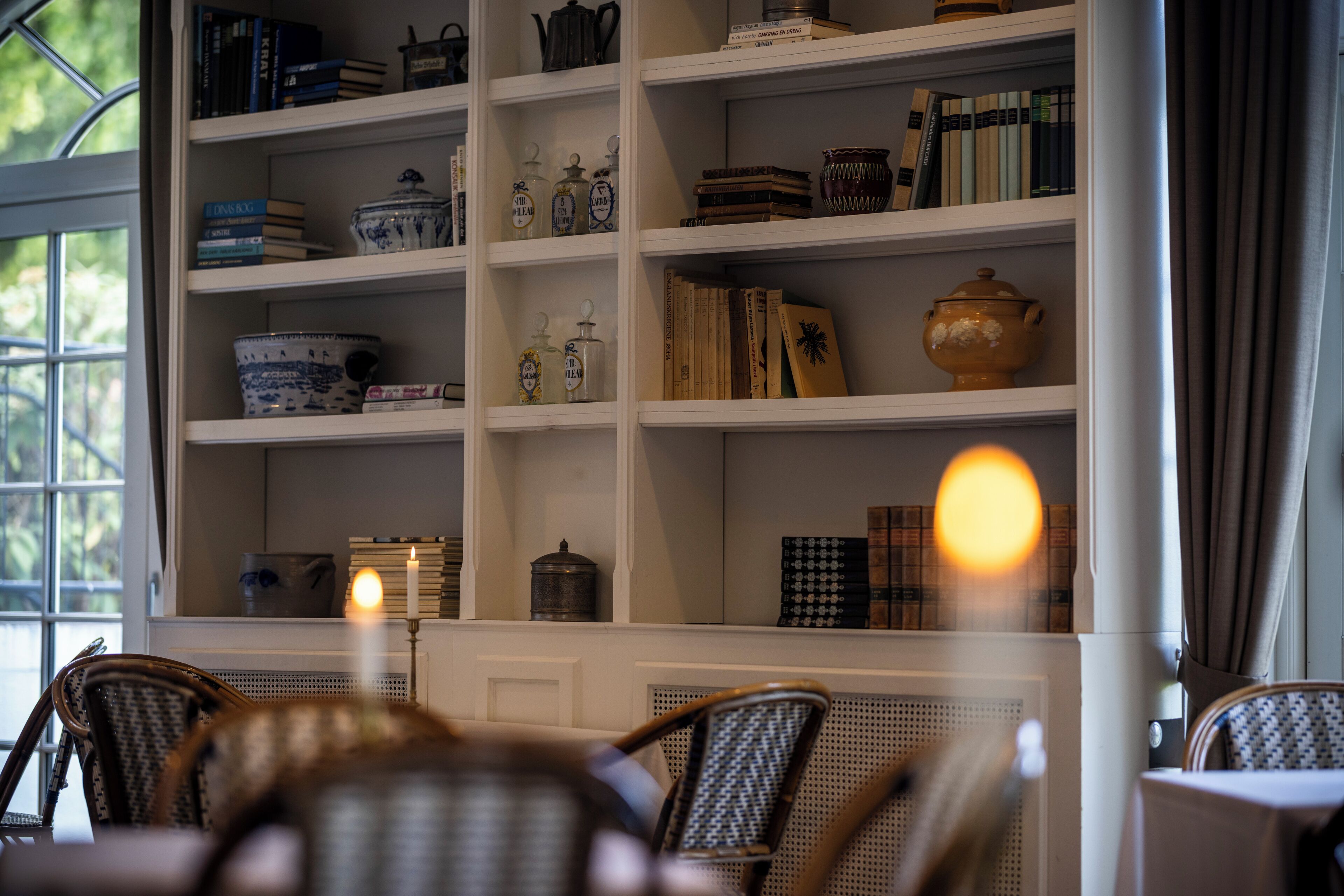 A room with a bookshelf filled with books and decorative items, a window with a view of greenery, and wicker chairs in the foreground.