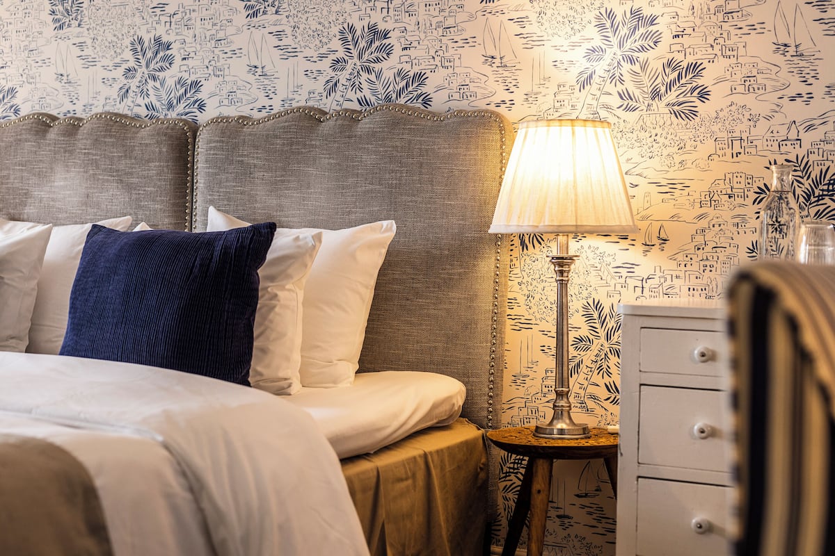 A neatly made bed with white and blue pillows, a lamp on a side table, and a patterned wallpaper.