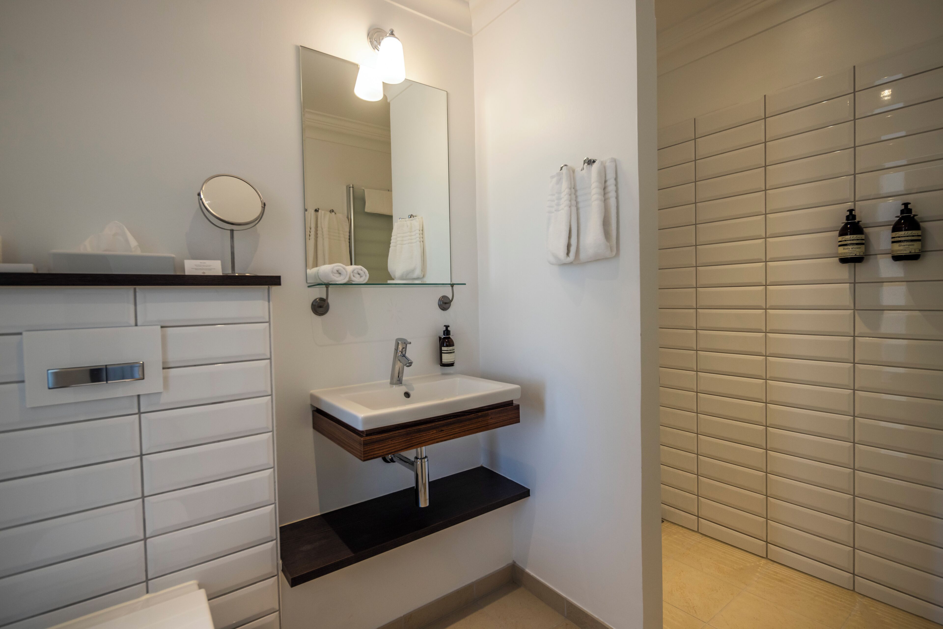 A modern bathroom with a white vanity, a round mirror, a glass shelf, a towel rack, and a shower area with tiled walls.