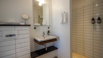 A modern bathroom with a white vanity, a round mirror, a glass shelf, a towel rack, and a shower area with tiled walls.
