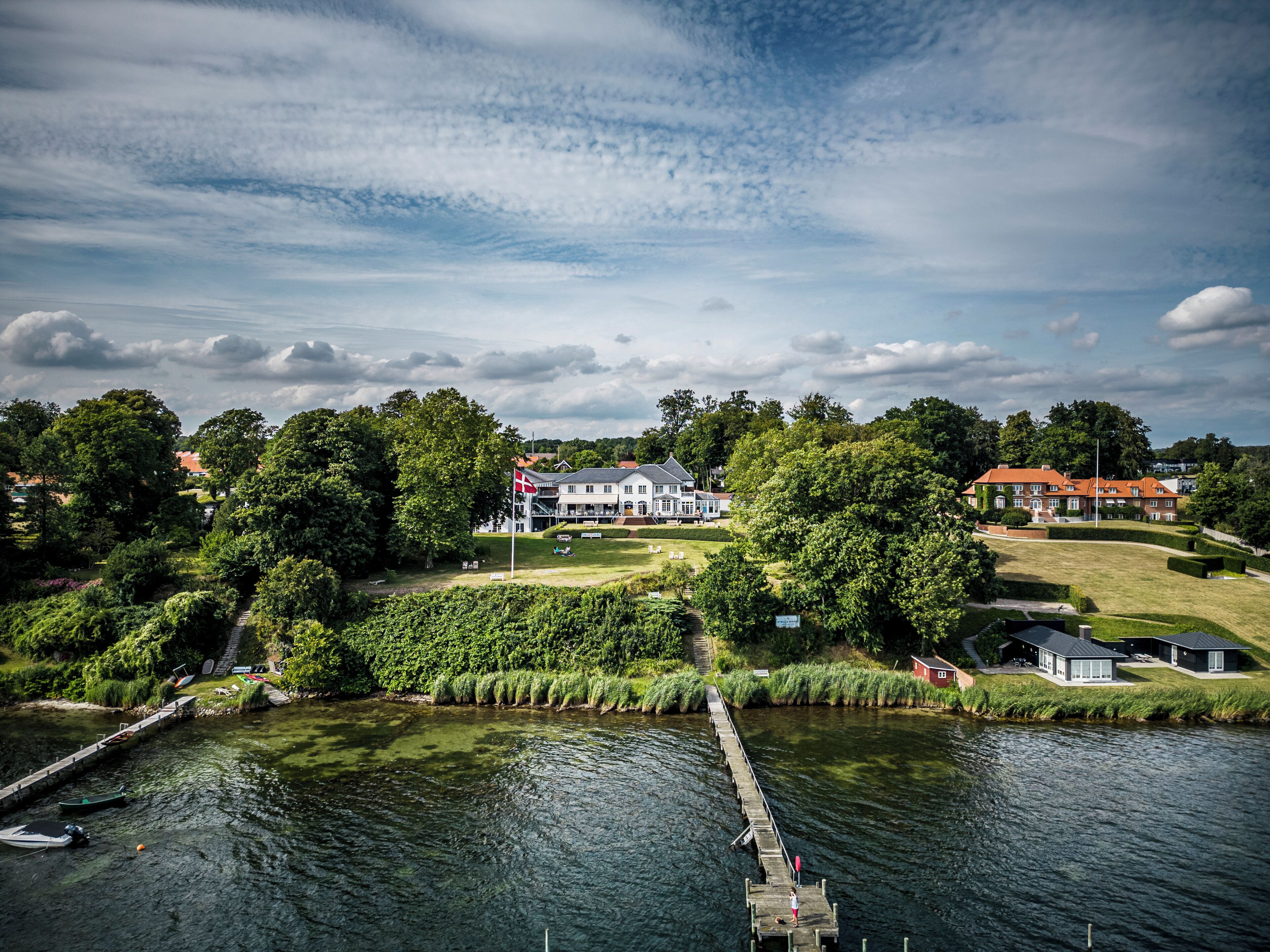 A riverside view with a dock extending into the water, surrounded by lush greenery and a large house with a red roof.