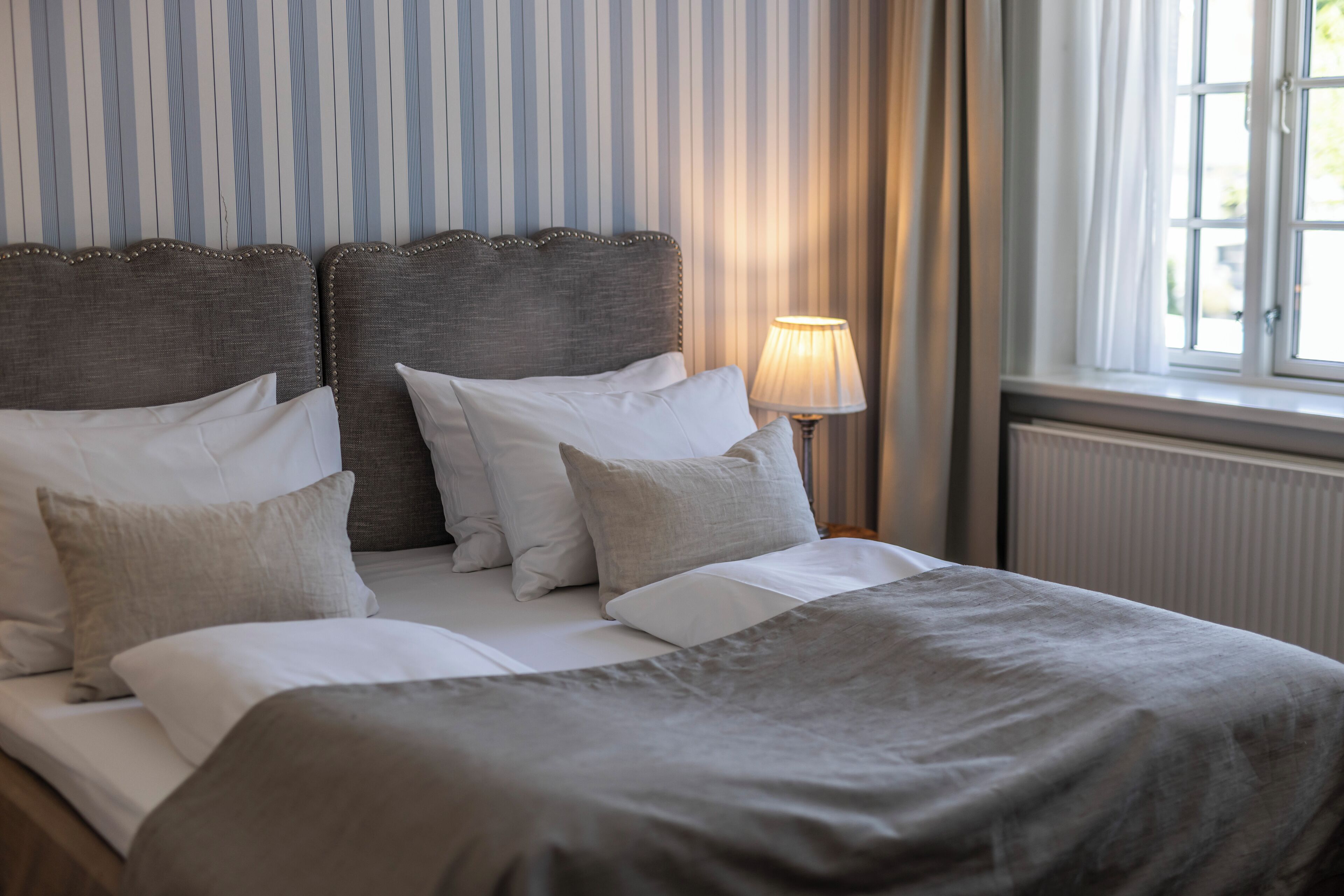 A double bed with a grey headboard and white bedding, a bedside lamp, and a window with curtains.