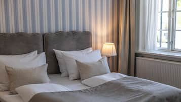 A double bed with a grey headboard and white bedding, a bedside lamp, and a window with curtains.