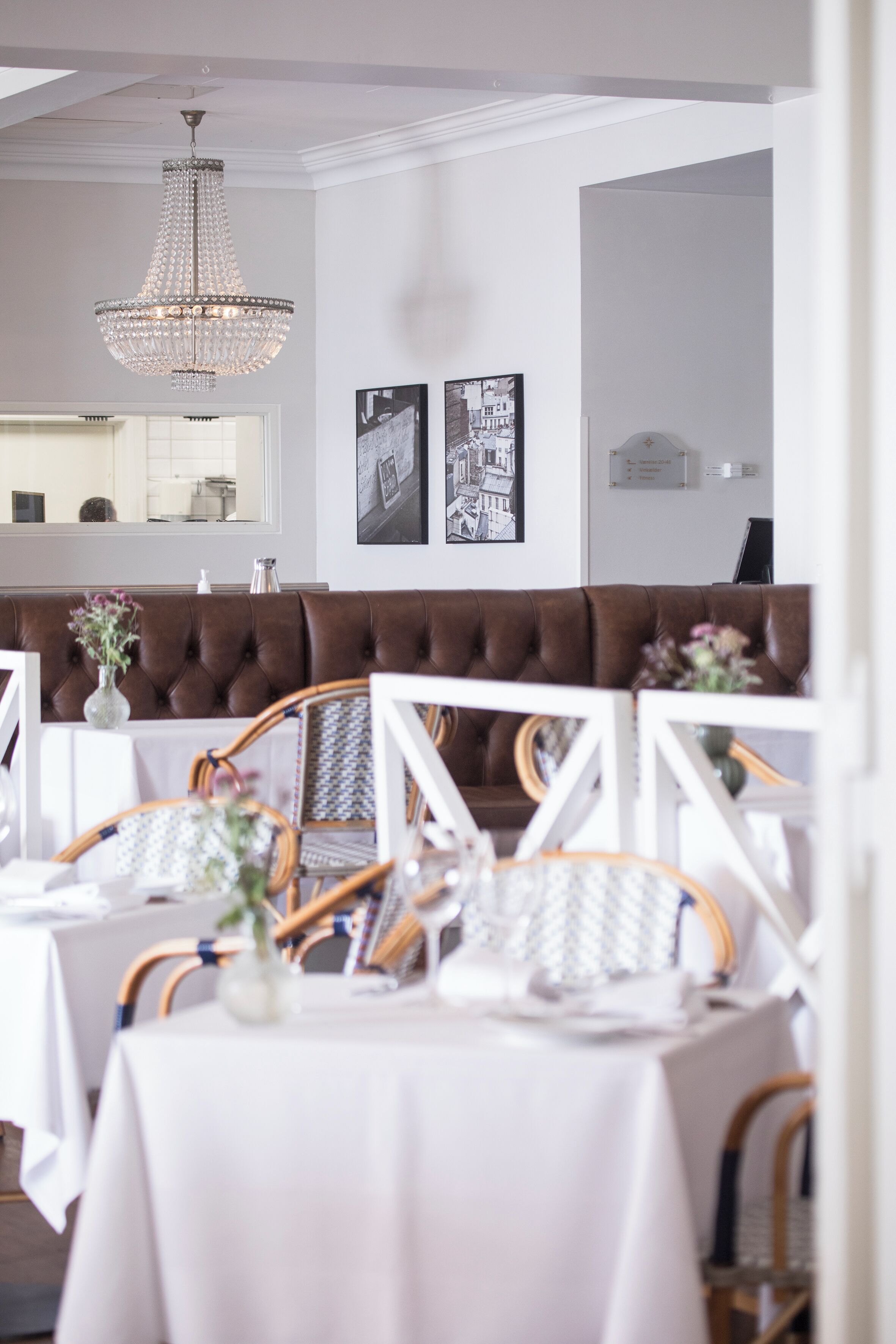 Un espace repas comprenant un banc recouvert d'un tissu marron, des tables blanches, des chaises en osier et un lustre.