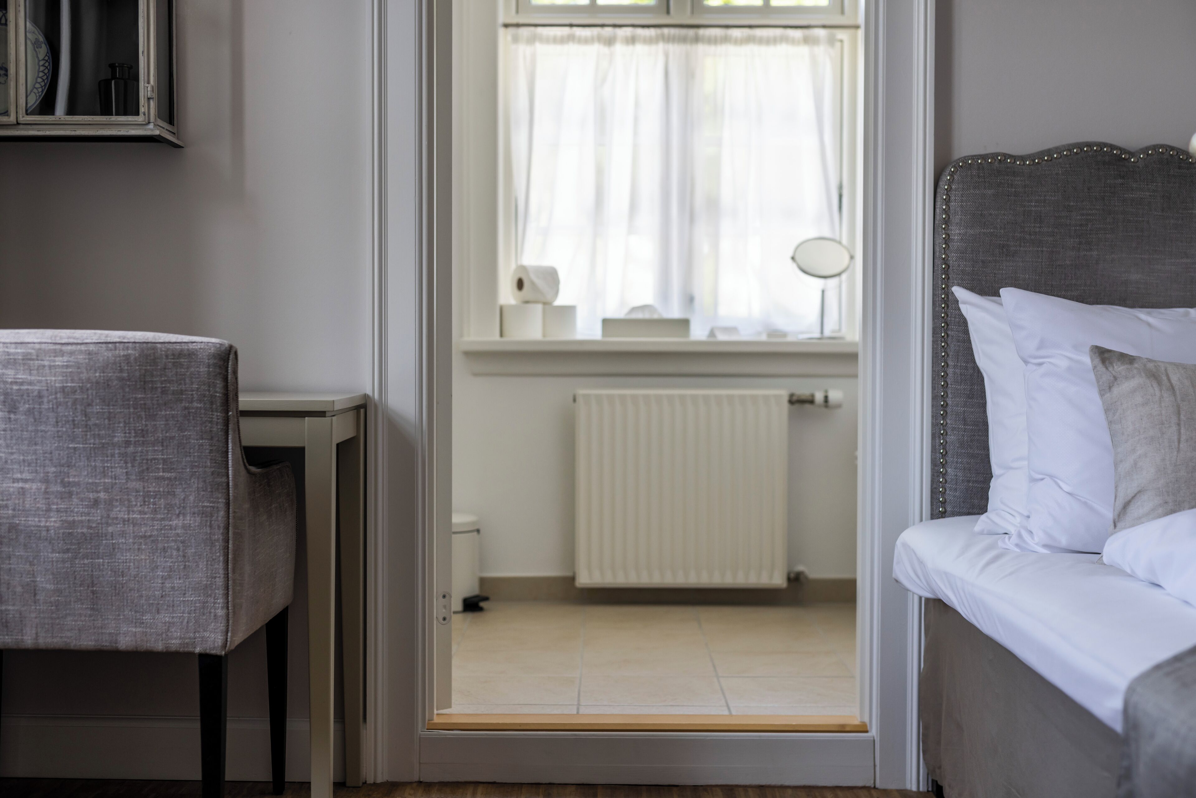 Una habitación con cama, una silla, una ventana con cortinas, un radiador y una mesita.