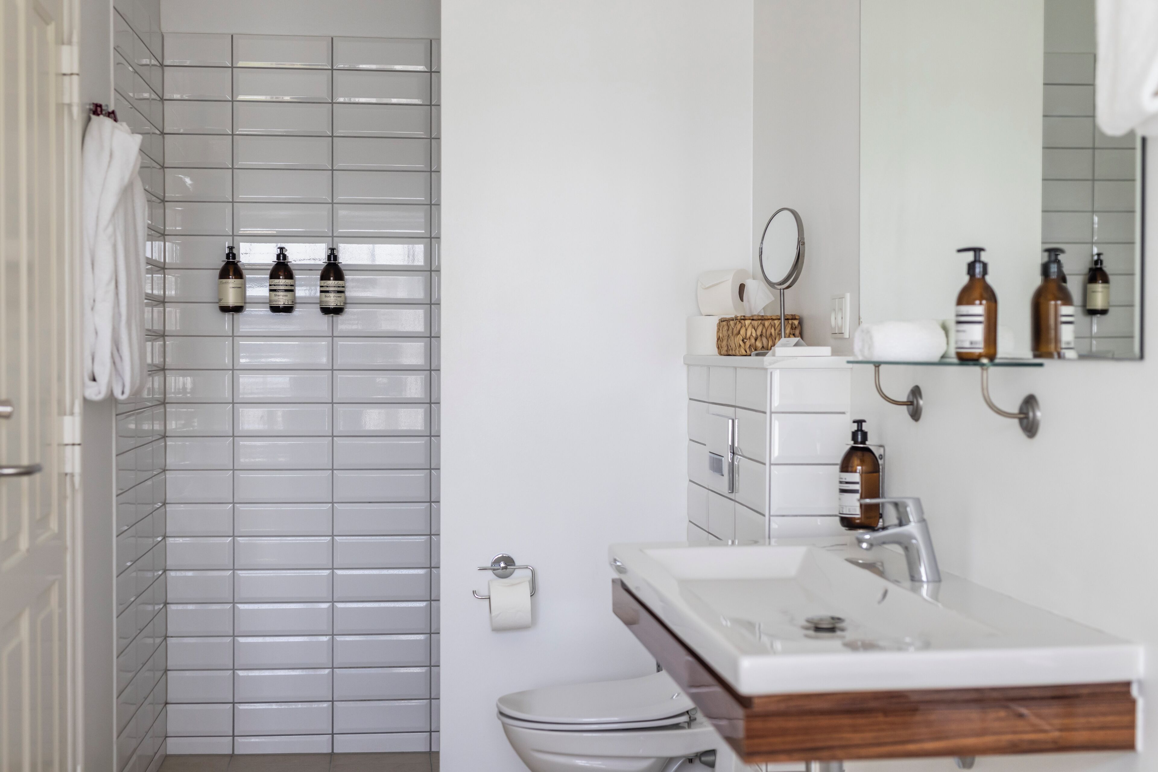 Un baño con paredes de azulejos blancos, un lavabo blanco con encimera de madera y artículos de aseo en una repisa.