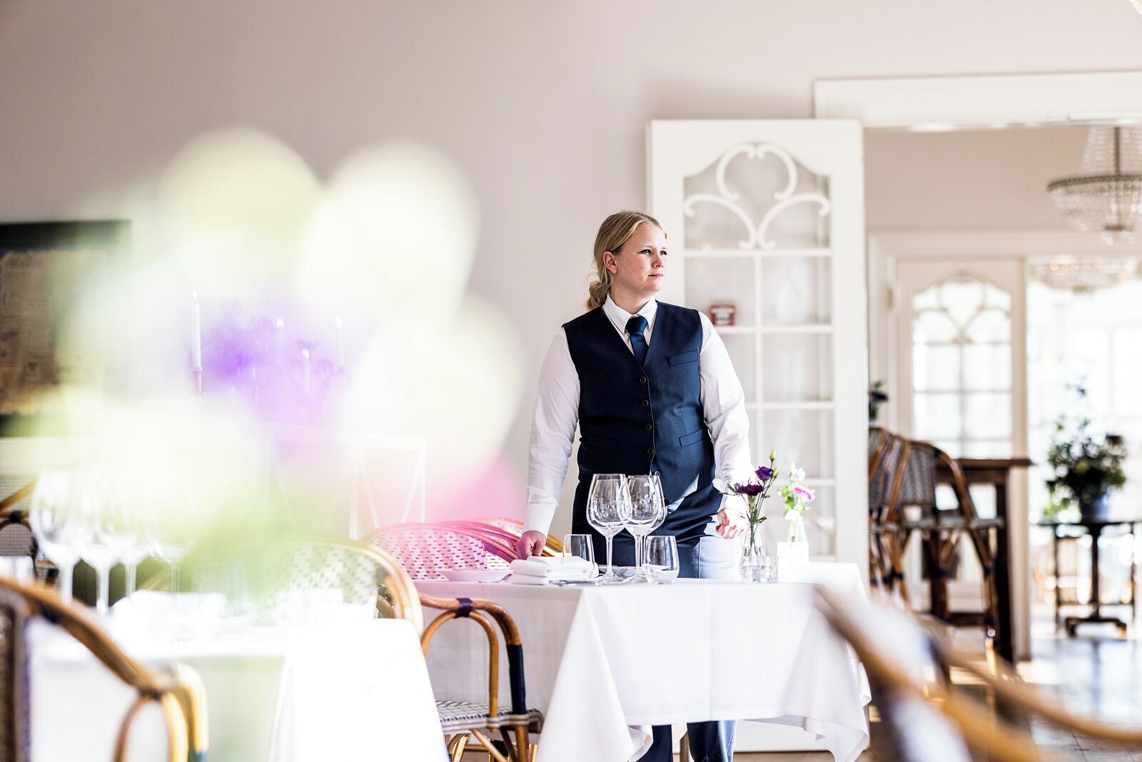 A dining area with a waiter standing by a table set with wine glasses and a vase of flowers.