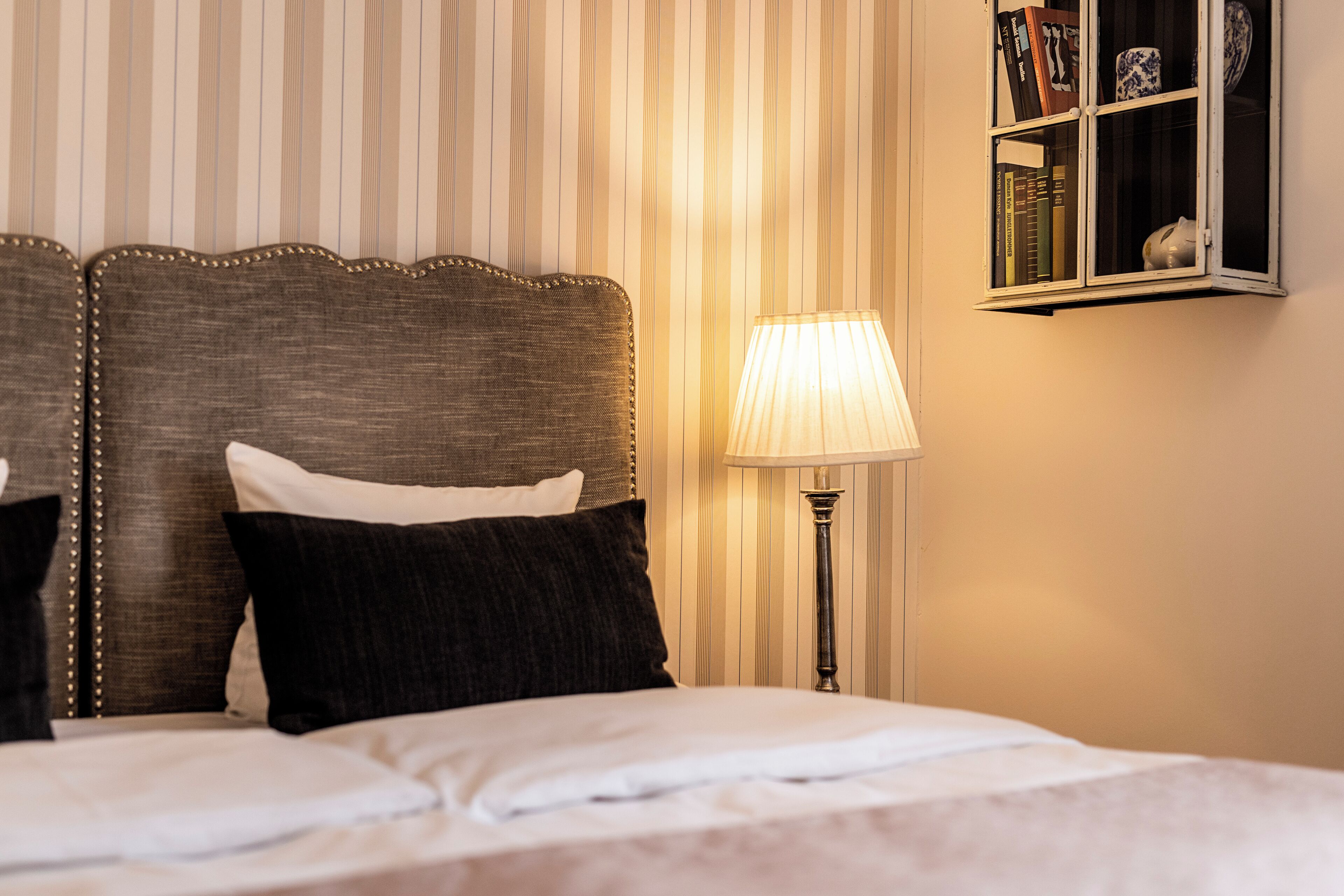 Una cama bien hecha con cabecera, una lámpara de noche y una estantería con libros y adornos.
