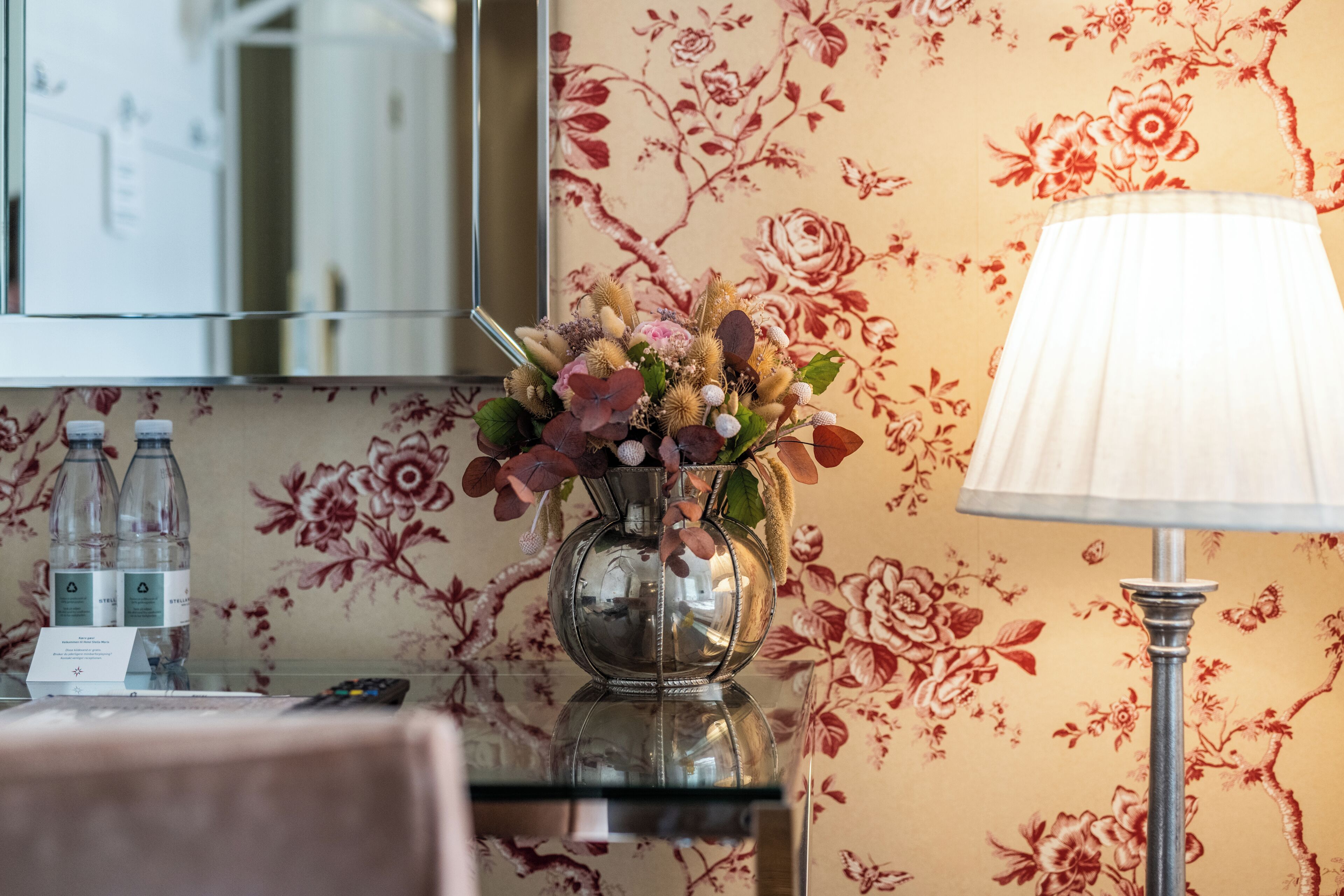 A hotel room with a floral wallpaper, a glass vase with flowers, a lamp, and two water bottles on a table.