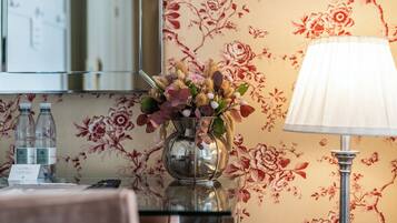 A hotel room with a floral wallpaper, a glass vase with flowers, a lamp, and two water bottles on a table.