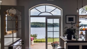 A room with a large arched window overlooking a lake, a wooden deck, and a view of a forested hill. - Stella Maris (Svendborg)