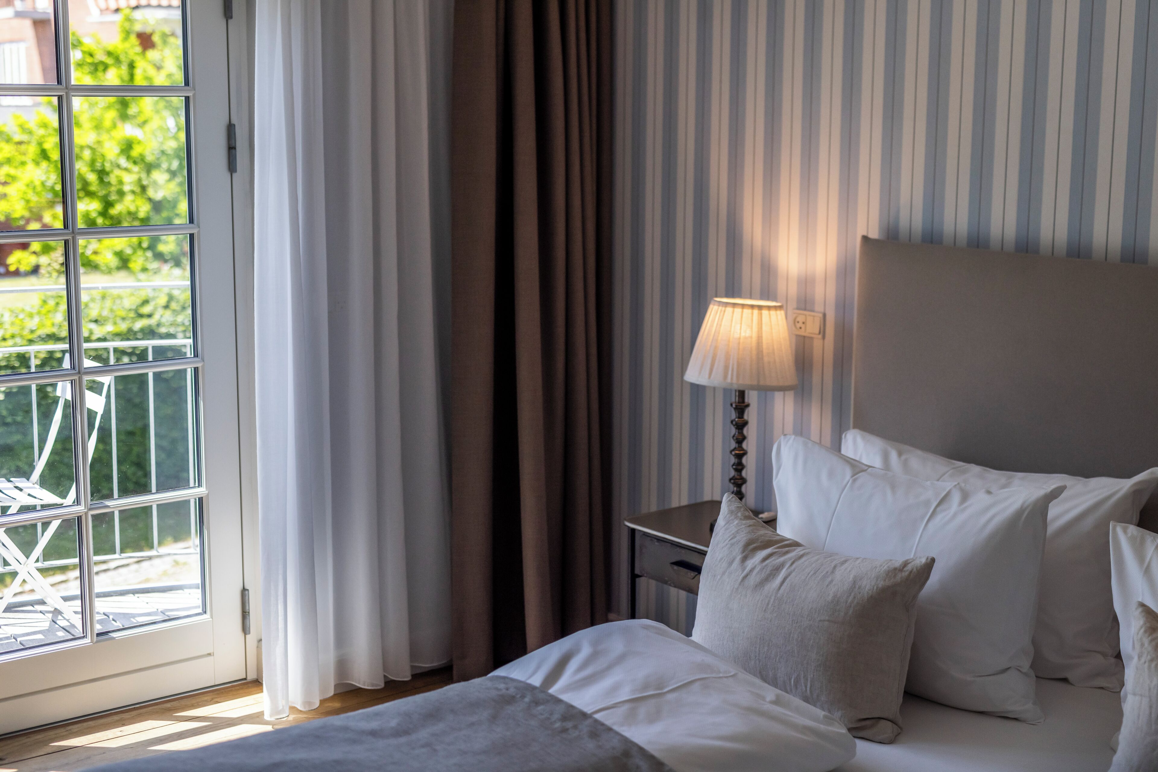 Habitación de hotel con cama, mesita de noche, lámpara y ventana con cortinas.