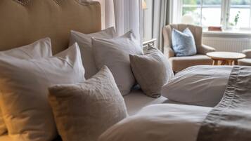 Una cama bien arreglada con varias almohadas y un cabecero, una mesita redonda pequeña y una silla en una habitación con ventana.