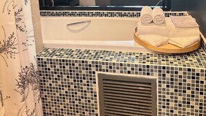 Een badkamer met een grote spiegel, een bad, een douchegedeelte en een raam met uitzicht op een slaapkamer.