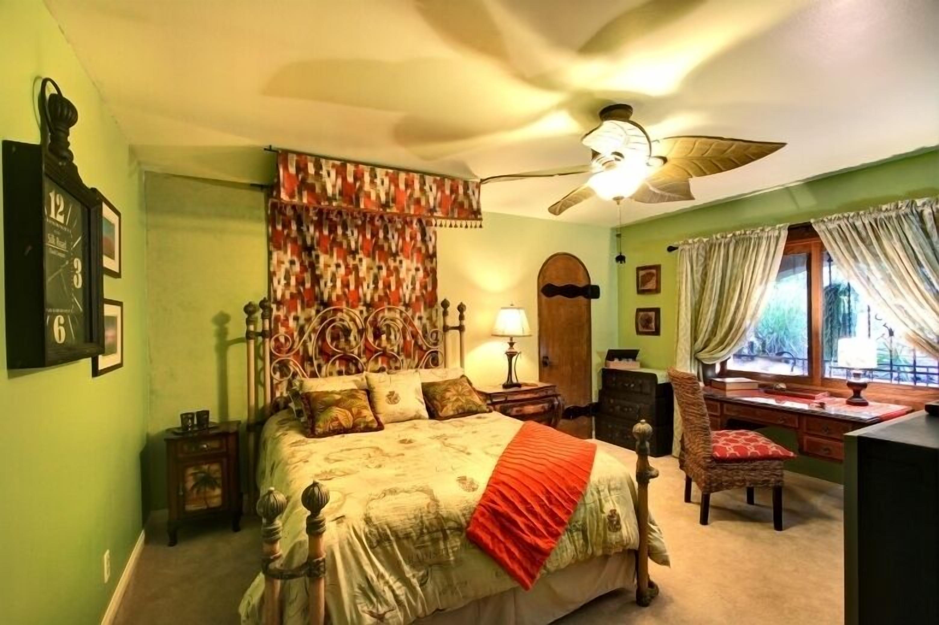 A bedroom with a bed, a desk, a chair, a ceiling fan, and a window with curtains.