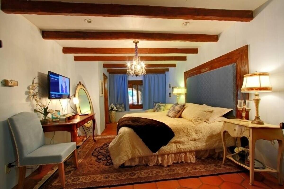 A bedroom with a large bed, wooden beams, a chandelier, a television, and a desk with a chair.