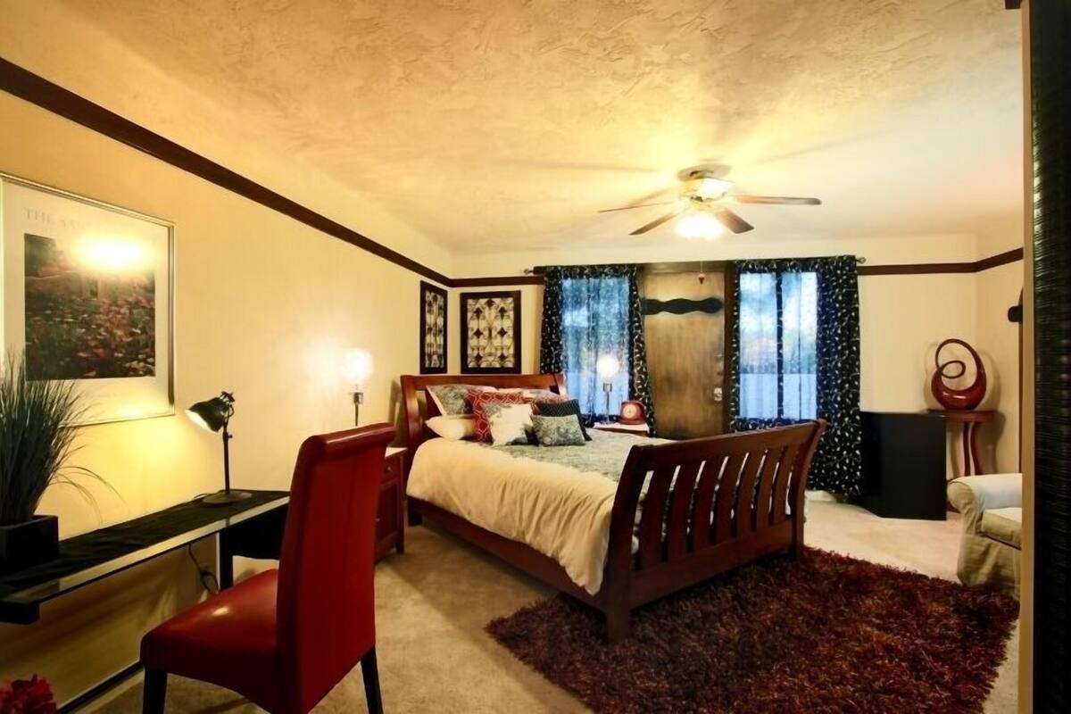 A bedroom with a bed, a desk, a chair, a ceiling fan, and a large window with curtains.