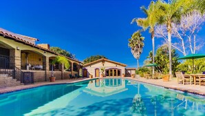 Una piscina cristalina rodeada de un patio con mobiliario exterior, un edificio con techo de tejas y palmeras.