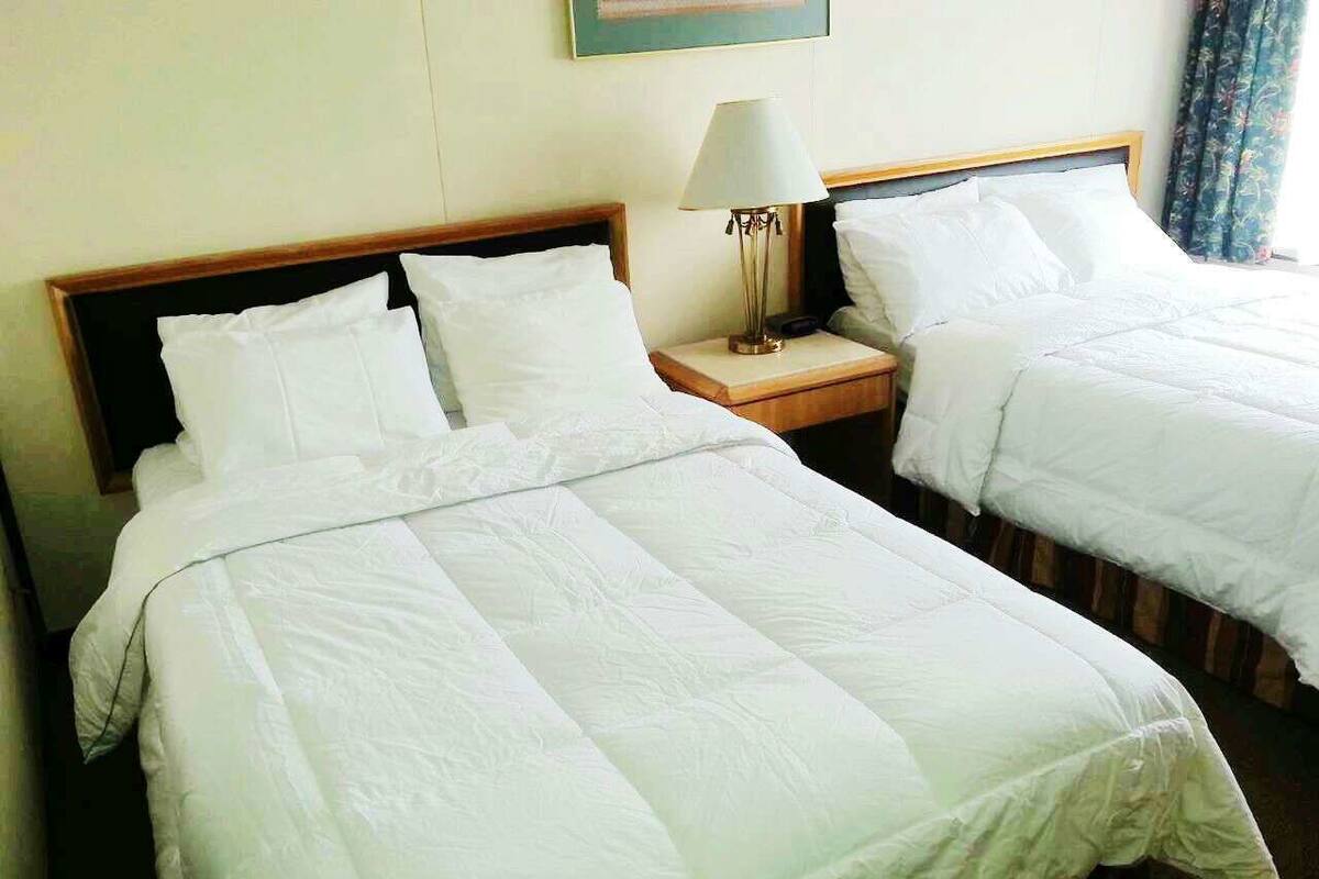 Habitación de hotel con dos camas individuales, cada una con ropa blanca y cabecero de madera. Hay una mesita de noche con lámpara entre las camas.