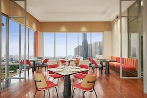 A modern dining area with large windows, red and blue chairs, and a city view. - Charm Suite Saigon (Ho Chi Minh City)