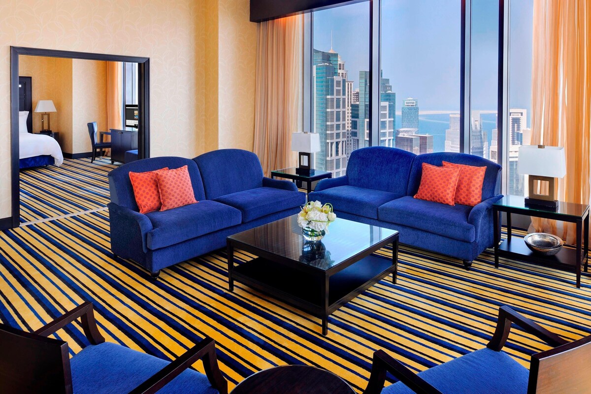 A hotel room with a large window offering a city view, two blue sofas with red cushions, a black coffee table, and a striped carpet.