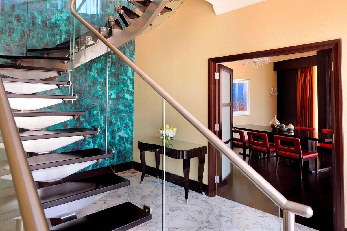 A modern staircase with glass railings and a dining area with red chairs and a table.