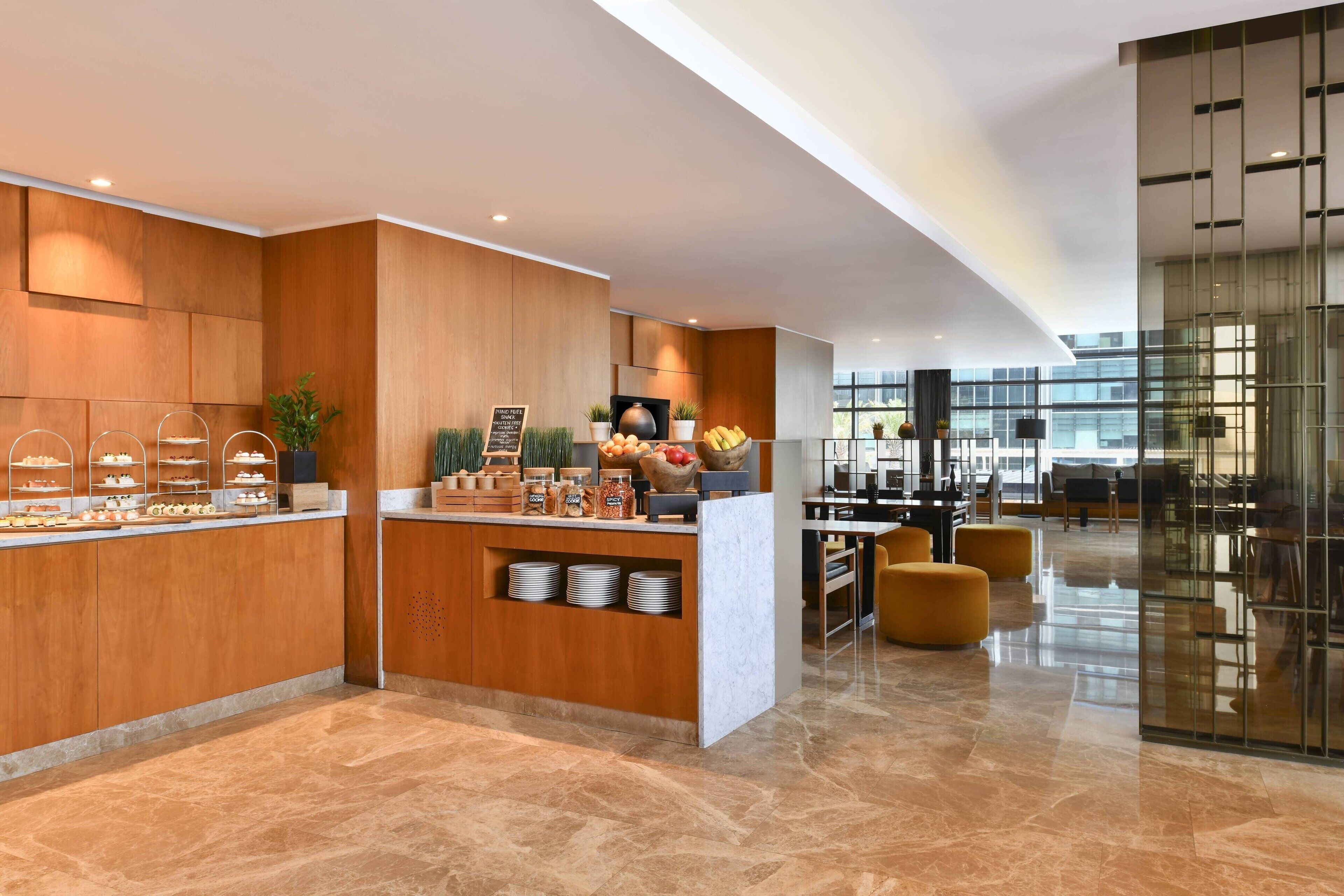 A modern hotel lobby with a wooden reception desk, a buffet area, and a seating area with yellow chairs.
