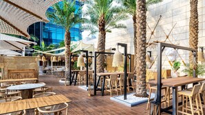 Un área de comedor al aire libre con mesas y sillas de madera, rodeada de palmeras y sombrillas.