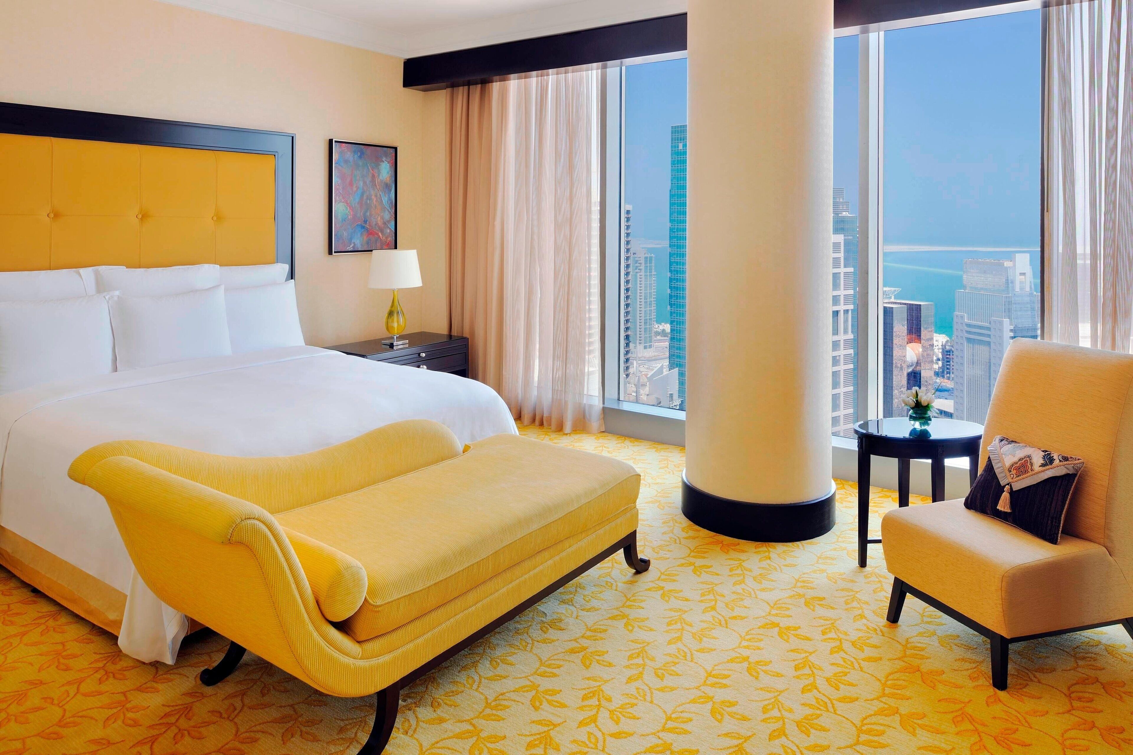 A hotel room with a large bed, a yellow sofa, a chair, a nightstand, a lamp, and a view of the city through large windows.