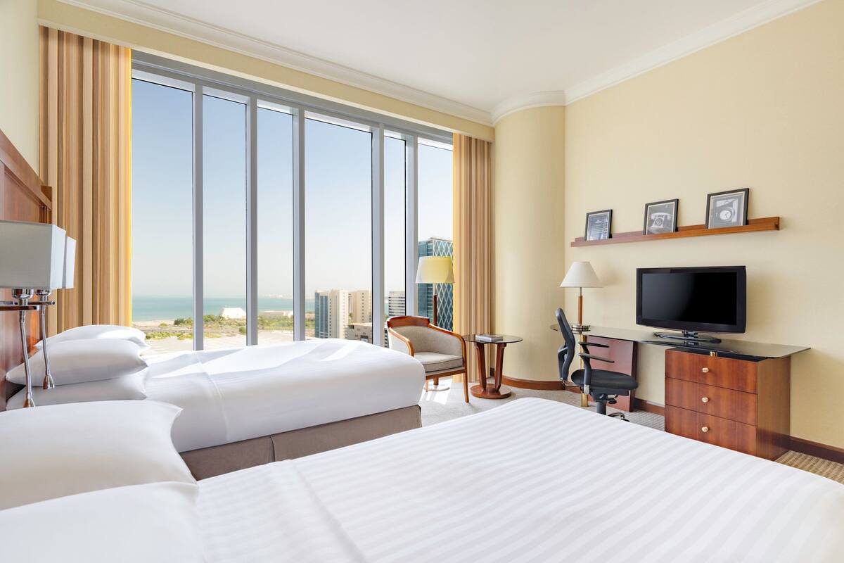 A hotel room with two beds, a desk, a chair, a television, and a large window with a view of the city and ocean.