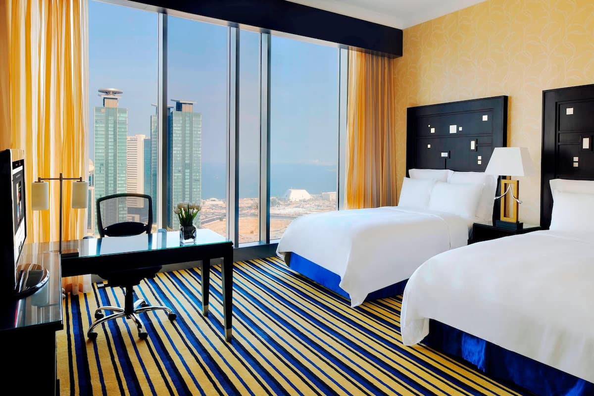 A hotel room with two beds, a desk, a chair, and a large window offering a city view.
