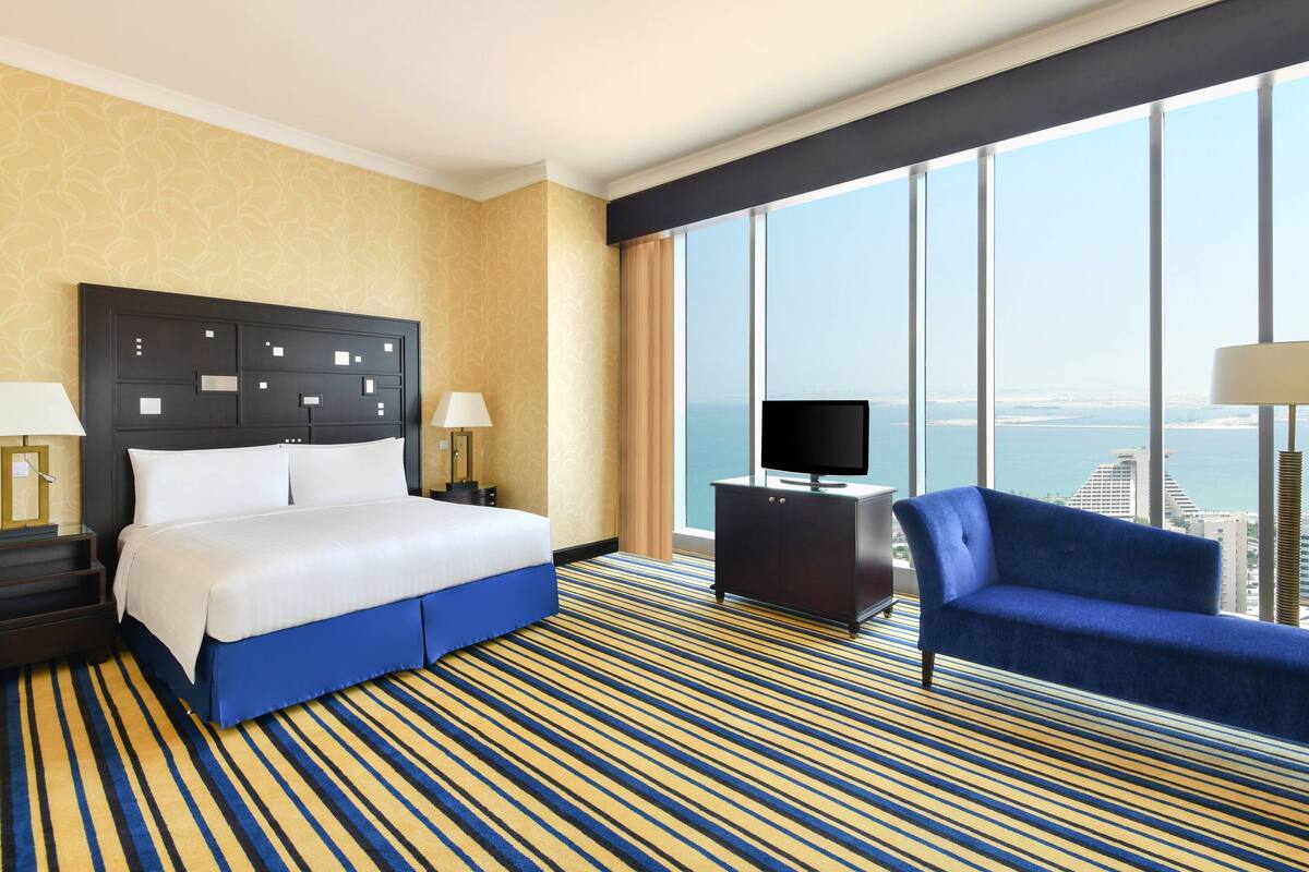 A hotel room with a large bed, a TV on a stand, a blue armchair, and a view of the city through large windows.